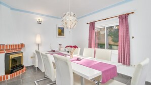 A dining area with a white table, chairs, a chandelier, a fireplace, and a window with curtains.