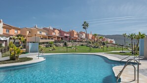 A swimming pool with a curved edge, surrounded by a stone wall, and adjacent to a row of multi-story buildings with balconies. - Holiday Home 'Bonita' with Sea Views, Shared Pool and Wi-Fi (ZAHARA DE LOS ATUNES)