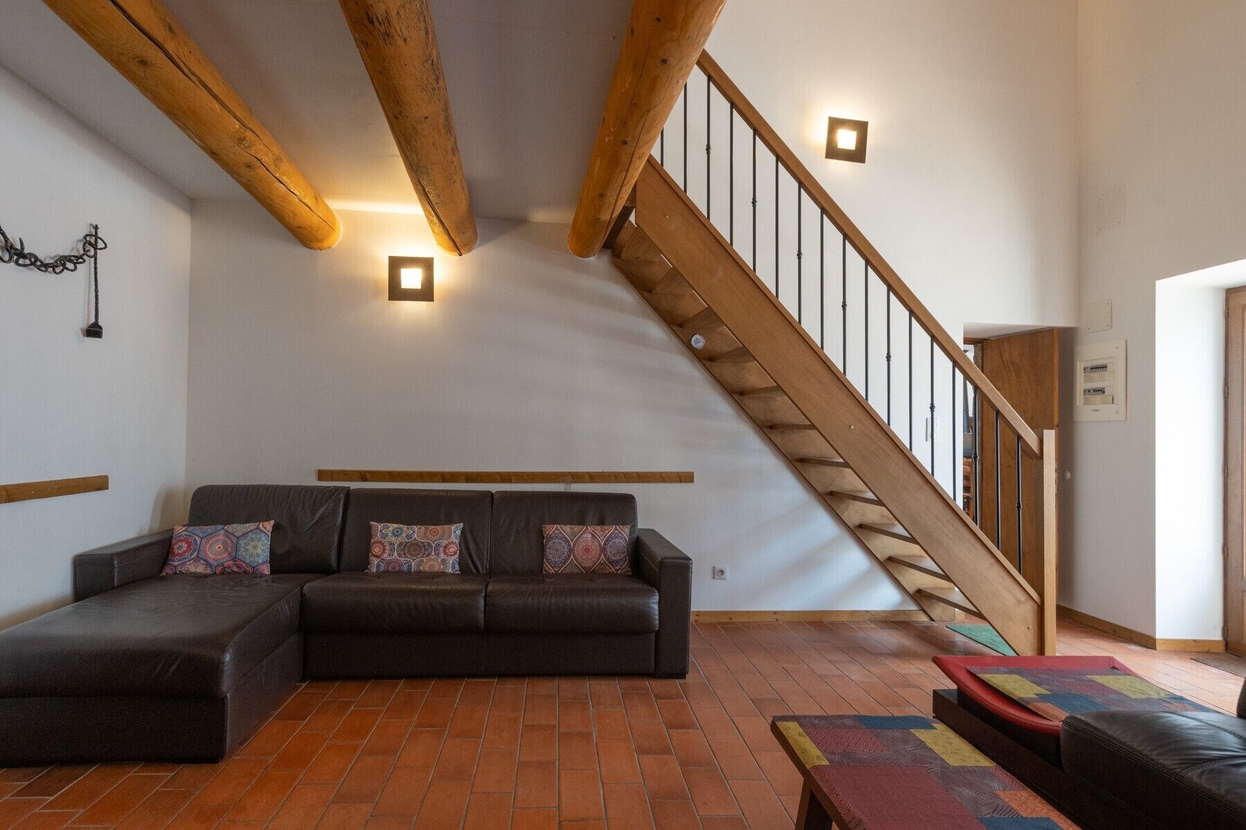 Un salon avec un canapé d’angle, un escalier en bois et des poutres apparentes.