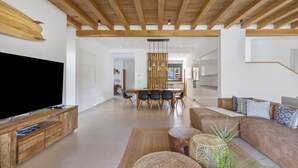 Ein modernes Wohnzimmer mit großem Flachbildfernseher, Holzbalkendecke, einem Essbereich mit Holztisch und -stühlen sowie einer gemütlichen Sitzecke mit Sofa und Zierkissen.