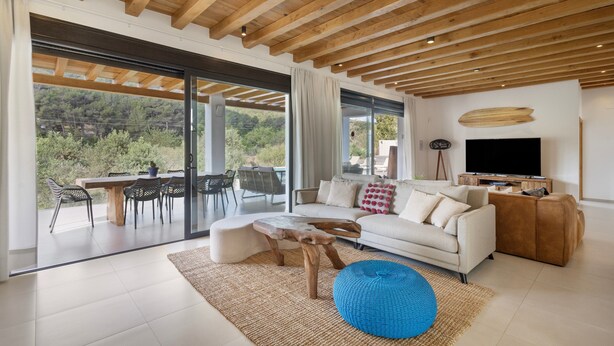Ein modernes Wohnzimmer mit Holzboden, einer Couch, einem Couchtisch, einem Fernseher und einem großen Fenster mit Blick auf Grünflächen.