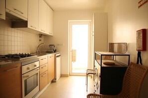 Una cocina con estufa, horno y armarios, un comedor con mesa y sillas, y una puerta que da al exterior.
