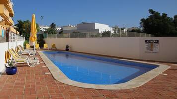 A swimming pool with lounge chairs and a sun umbrella, surrounded by a brick patio and a white wall with a sign.