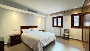 Un dormitorio con cama, mesitas de noche, radiador, ventana, una silla y un armario.