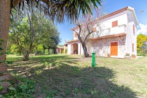 A building with a stone facade, wooden shutters, and a garden with trees and a green post.