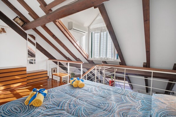 Une pièce avec un plafond en pente, des poutres en bois, un lit avec des serviettes roulées, un escalier et une fenêtre avec des rideaux.