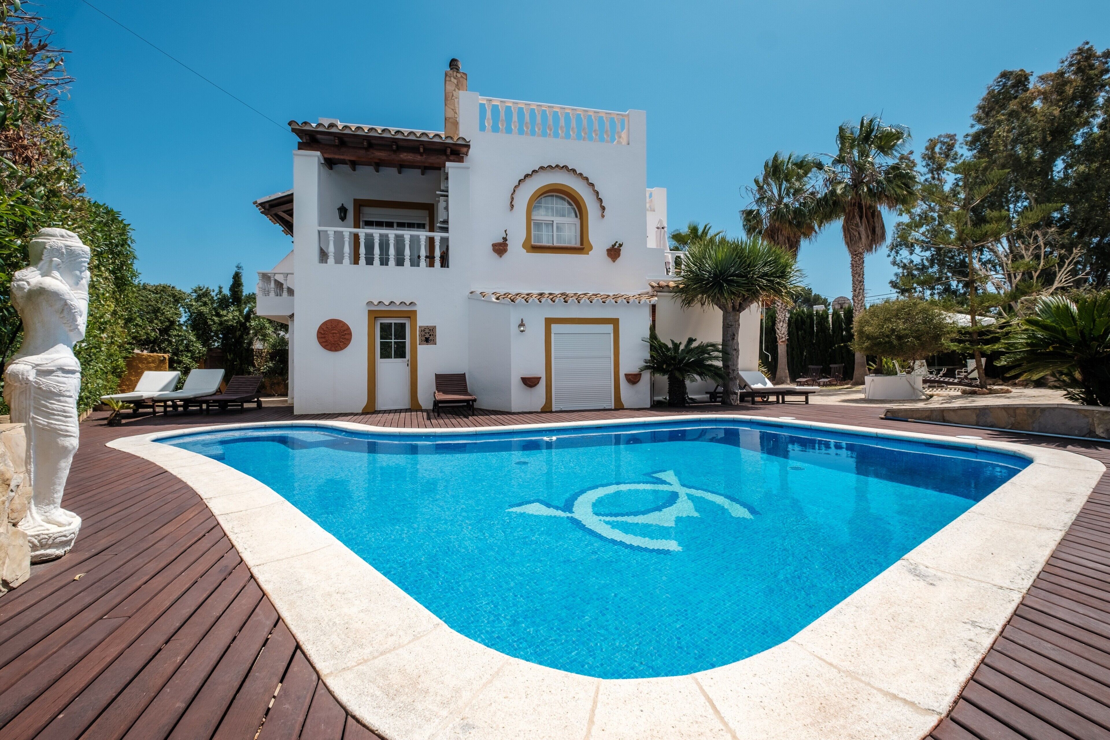 Una piscina con el símbolo de socorrista, un edificio blanco con balcones y una estatua.