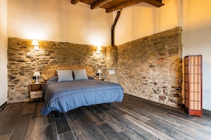 A bedroom with a stone wall, a bed with a blue bedspread, two bedside lamps, and a wooden nightstand.
