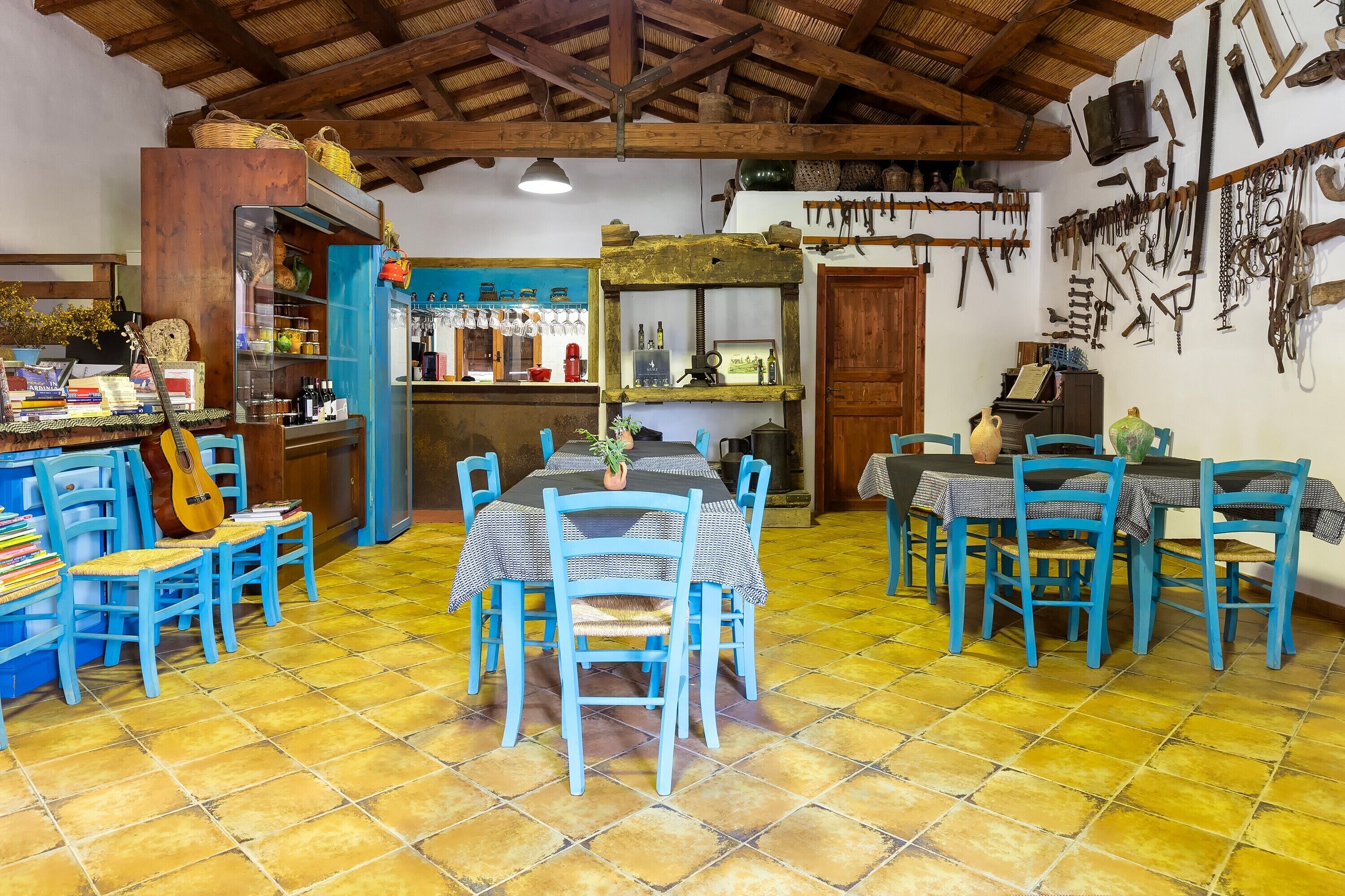 Un coin repas rustique avec des chaises bleues et une table, un bar avec des étagères remplies de bouteilles, et un mur où sont accrochés des outils.