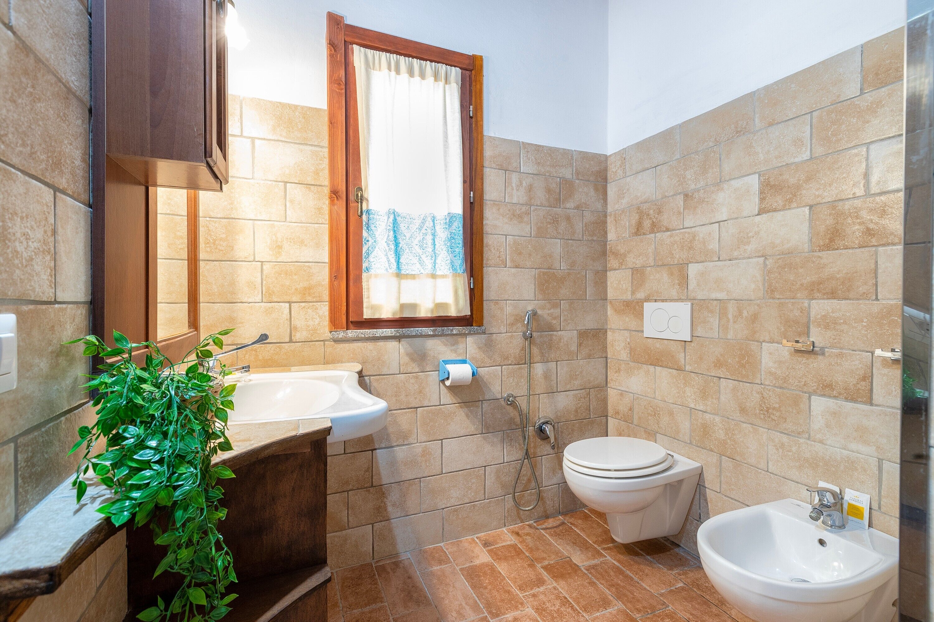 A bathroom with a sink, toilet, bidet, and a window with a curtain.