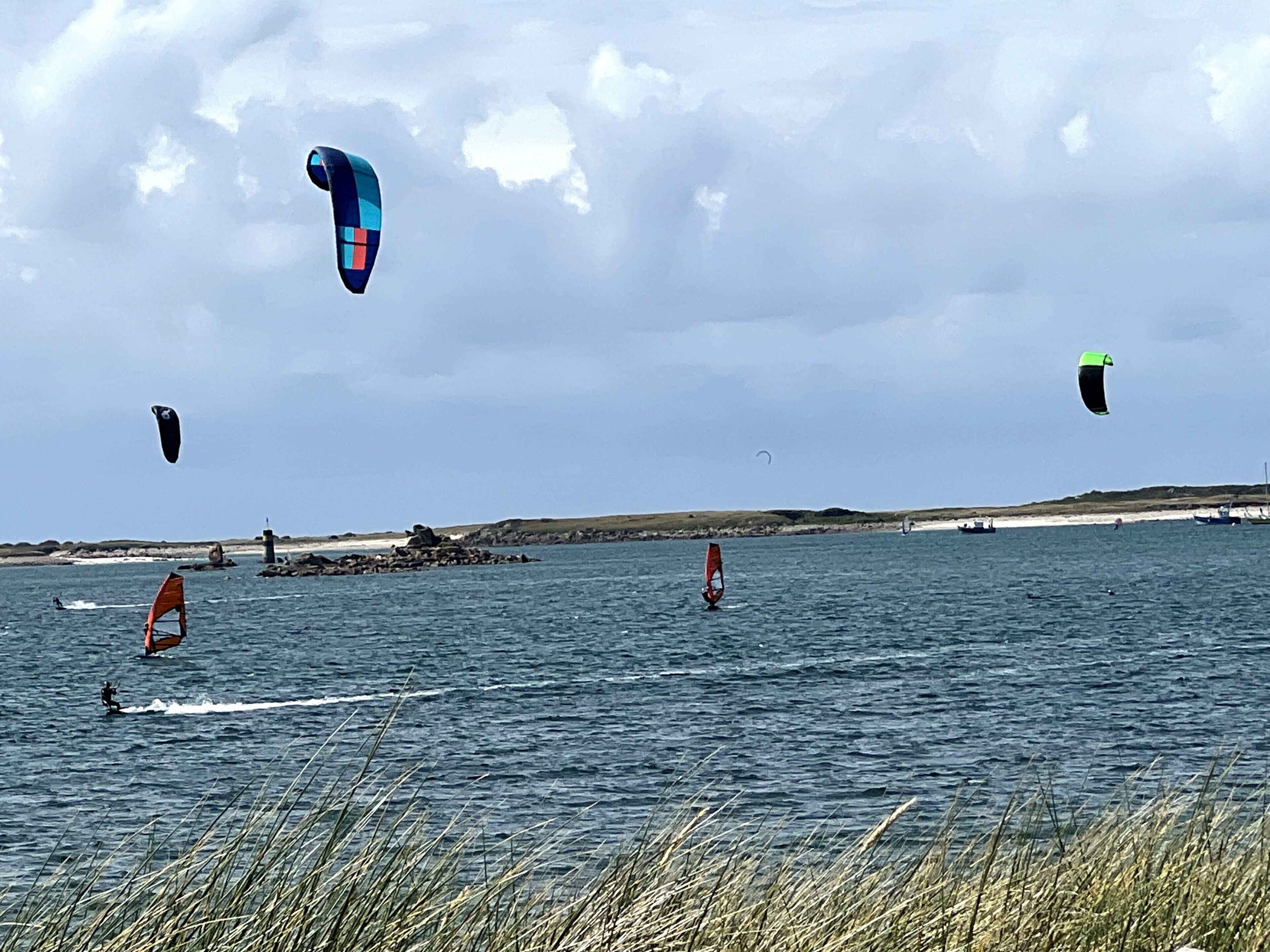 Escena costera con personas practicando windsurf, mar tranquilo y costa rocosa en la distancia.