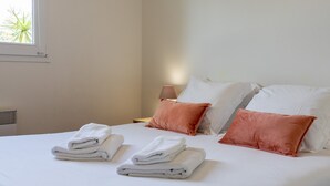 A neatly made bed with white and peach pillows, a lamp, and folded towels on a white bedspread.