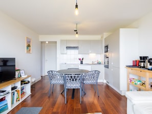 A modern kitchen with white cabinets, a dining table with four chairs, a television on a stand, and a bookshelf with books and a picture frame.