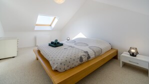 Une chambre avec un plafond en pente, un puits de lumière, un lit avec une couette à motifs pois et décorée de vélos, une table de chevet avec une lampe et une commode blanche.