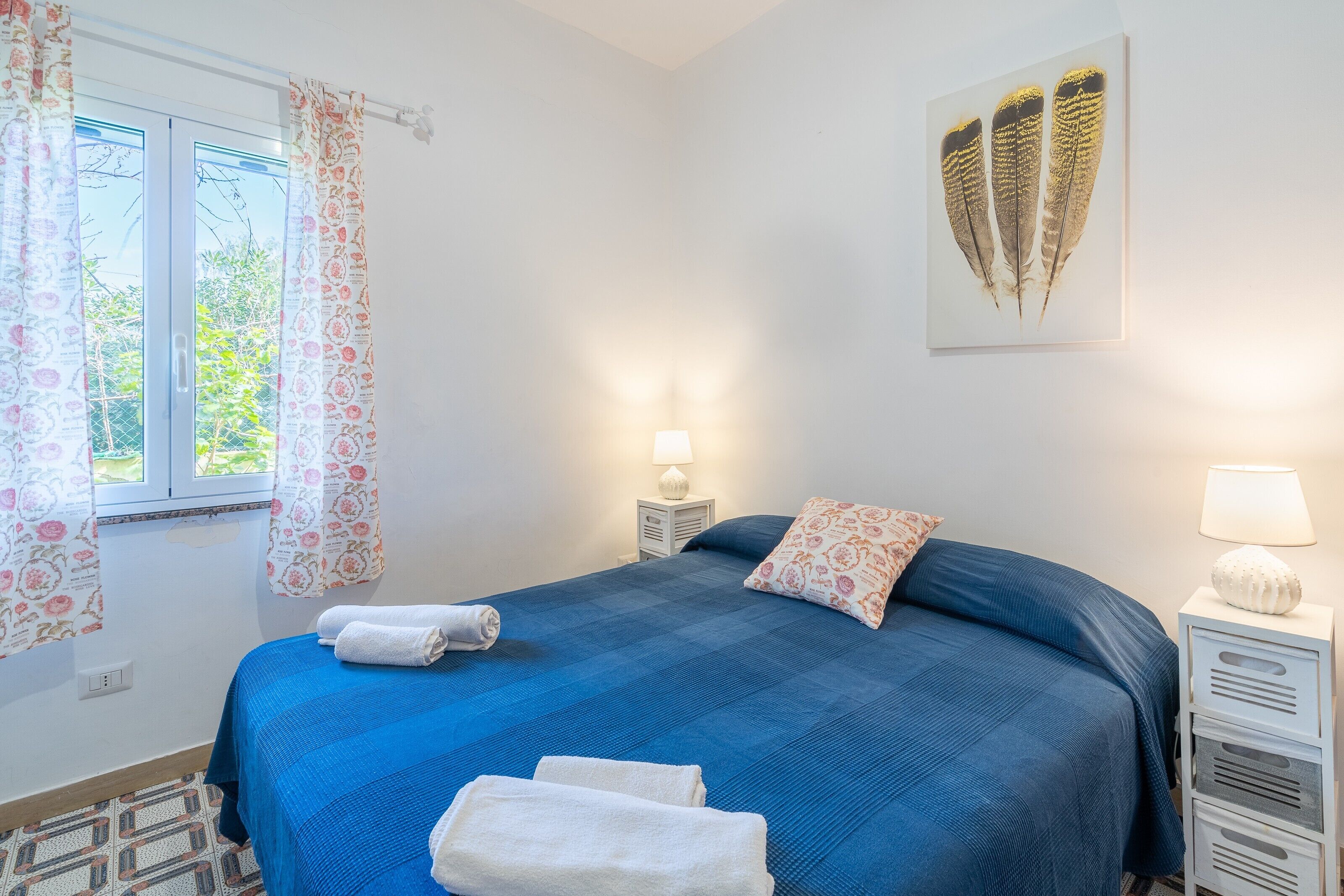 Une chambre avec une couvre-lit bleue, deux oreillers, une œuvre d’art accrochée au mur, une fenêtre avec des rideaux et une lampe de chevet.