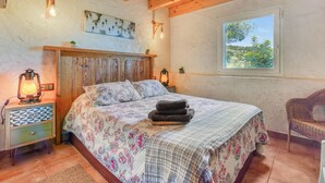 A bedroom with a wooden headboard, a bed with floral bedding, a nightstand with a lamp, and a window with a view of greenery. - Country House 'El Castellet De L'Ocaive' with Mountain View, Wi-Fi and Air Conditioning (Alicante)