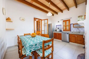 Un comedor con una mesa dispuesta para cuatro, sillas de madera, un frutero y una cocina al fondo con refrigerador, microondas y armarios de madera.