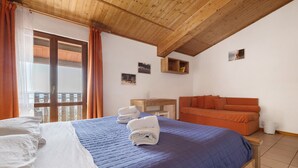 A bedroom with a bed, a wooden headboard, a wooden desk, a sofa, and a balcony with curtains. - Room 'Camera Familiare Classic' with Lake View, Shared Garden and Wi-Fi (Toscolano Maderno)