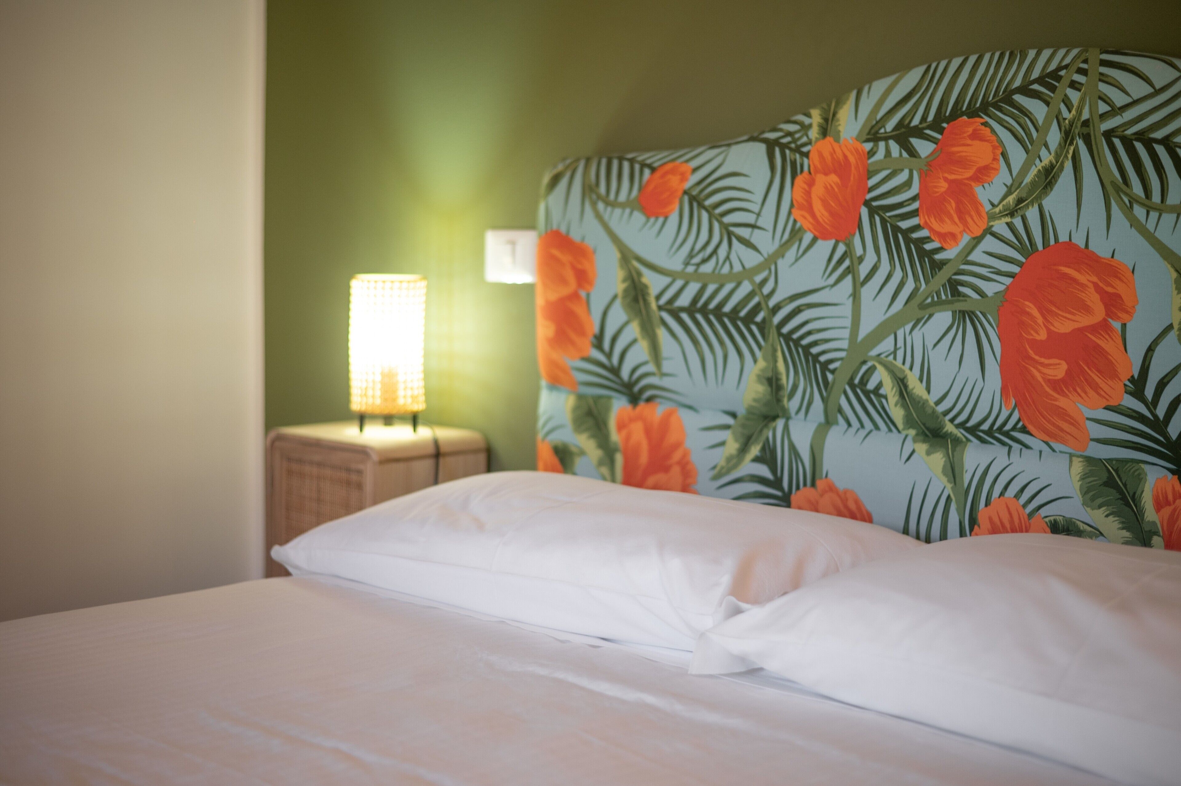 Una cama con ropa de cama blanca, un cabecero con estampado floral, una mesita de noche con lámpara y una pared verde.