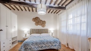 A bedroom with a bed, white wardrobe, wooden beams, a wall-mounted decorative piece, and a window with sheer curtains. - Apartment 'Pendolino' with Shared Pool, Wi-Fi and Air Conditioning (Capalbio)
