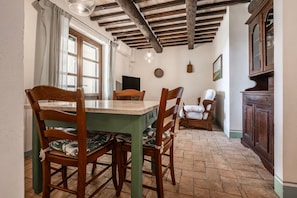 Uma área de refeições com mesa e cadeiras de madeira, um banco, um armário e uma janela com cortinas.
