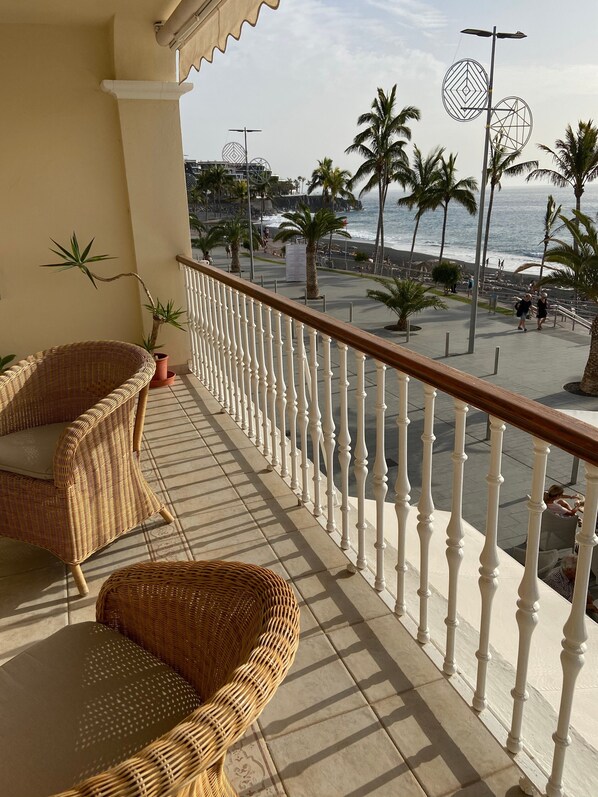 Un balcón con mobiliario de mimbre, barandilla blanca y vistas a la playa y palmeras.