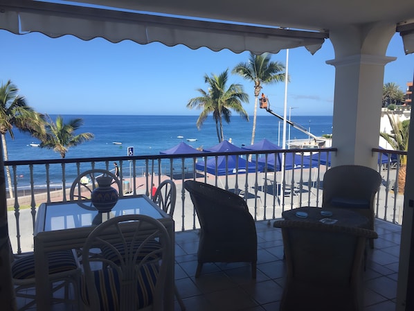 Un balcon avec des chaises et une table, d’où l’on peut admirer la mer, les palmiers et un panorama sur l’océan.