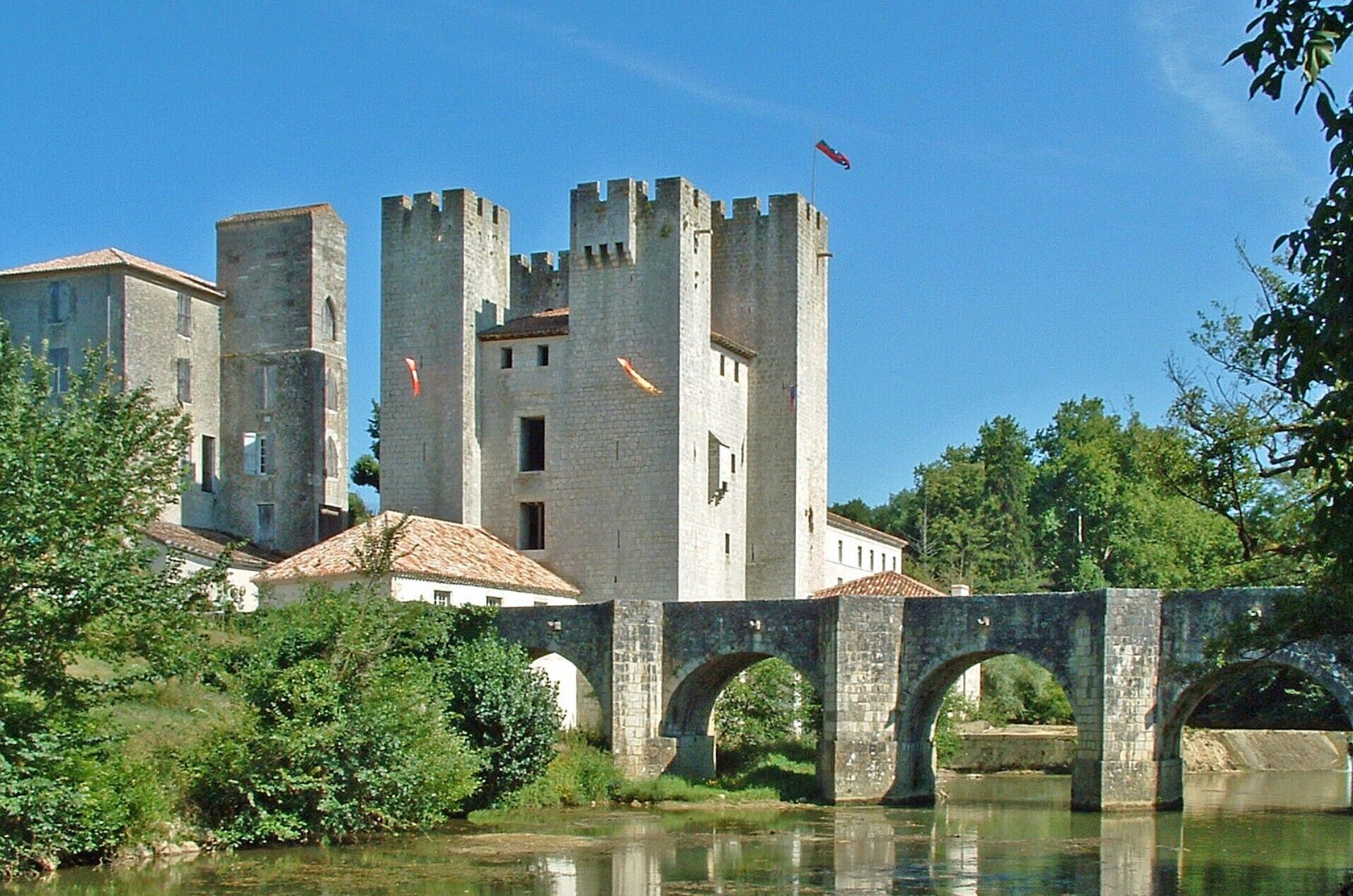 Een historisch stenen kasteel met meerdere torens, een brug en weelderige begroeiing.