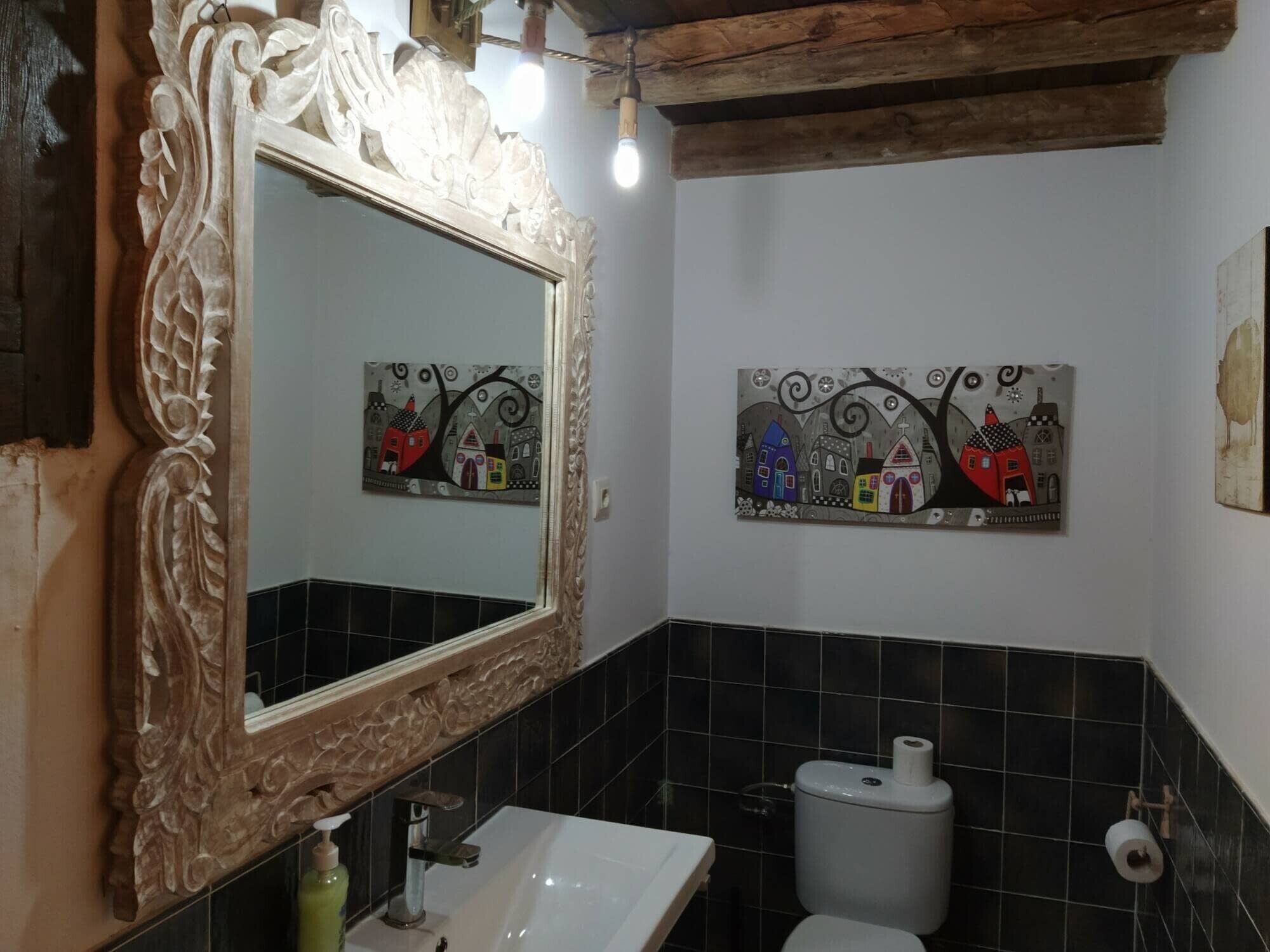 A bathroom with a large ornate mirror, a white sink, a toilet, and two wall-mounted artworks depicting stylized houses.