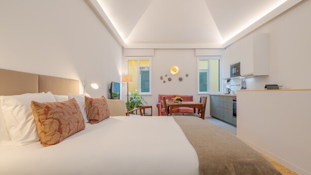 Un dormitorio compacto con cama, mesita de noche, lámpara y una pequeña zona de kitchenette.