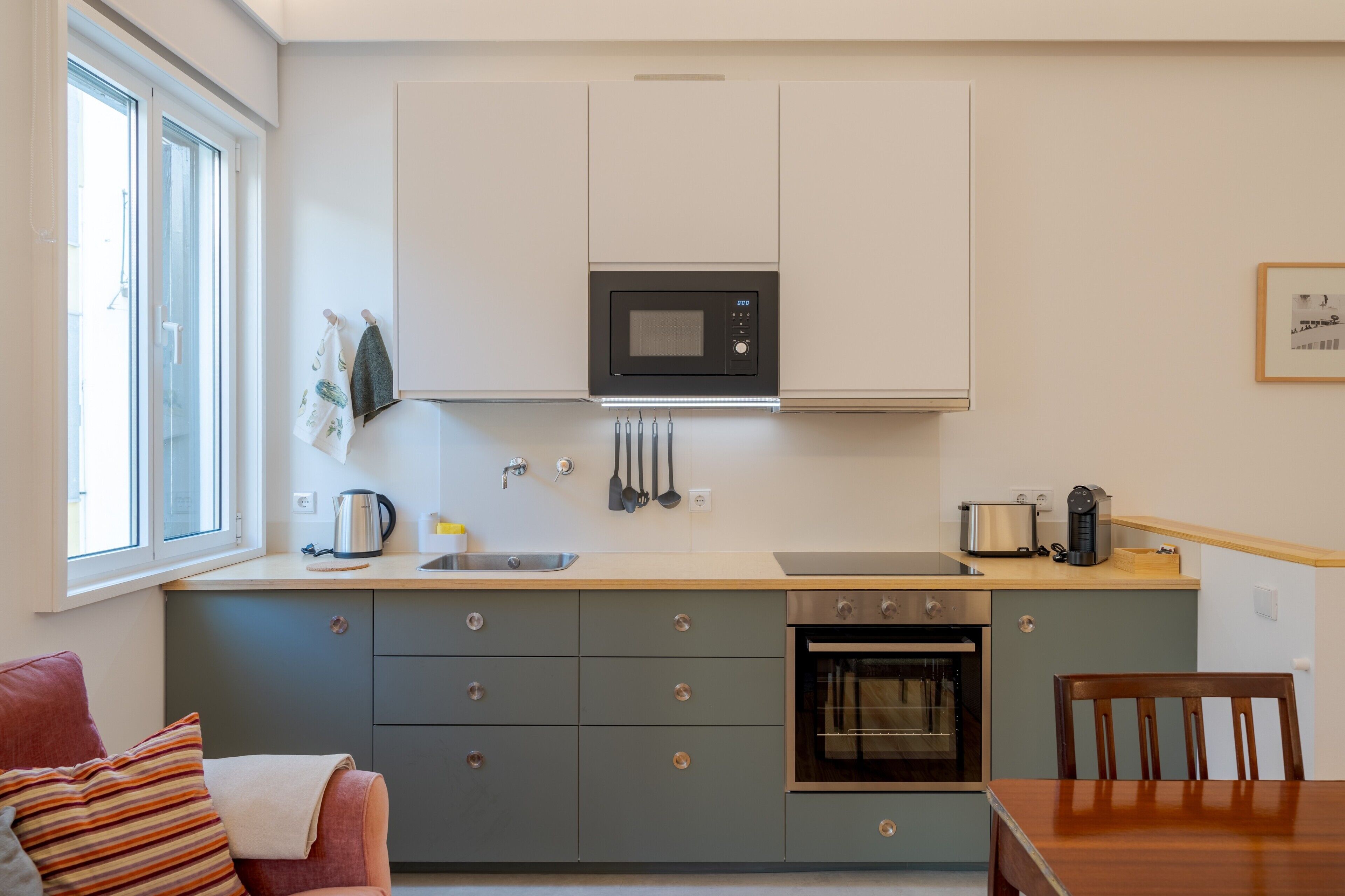 Een praktische keuken met een magnetron, oven en spoelbak, een eettafel met stoelen, en een raam dat voor natuurlijk licht zorgt.
