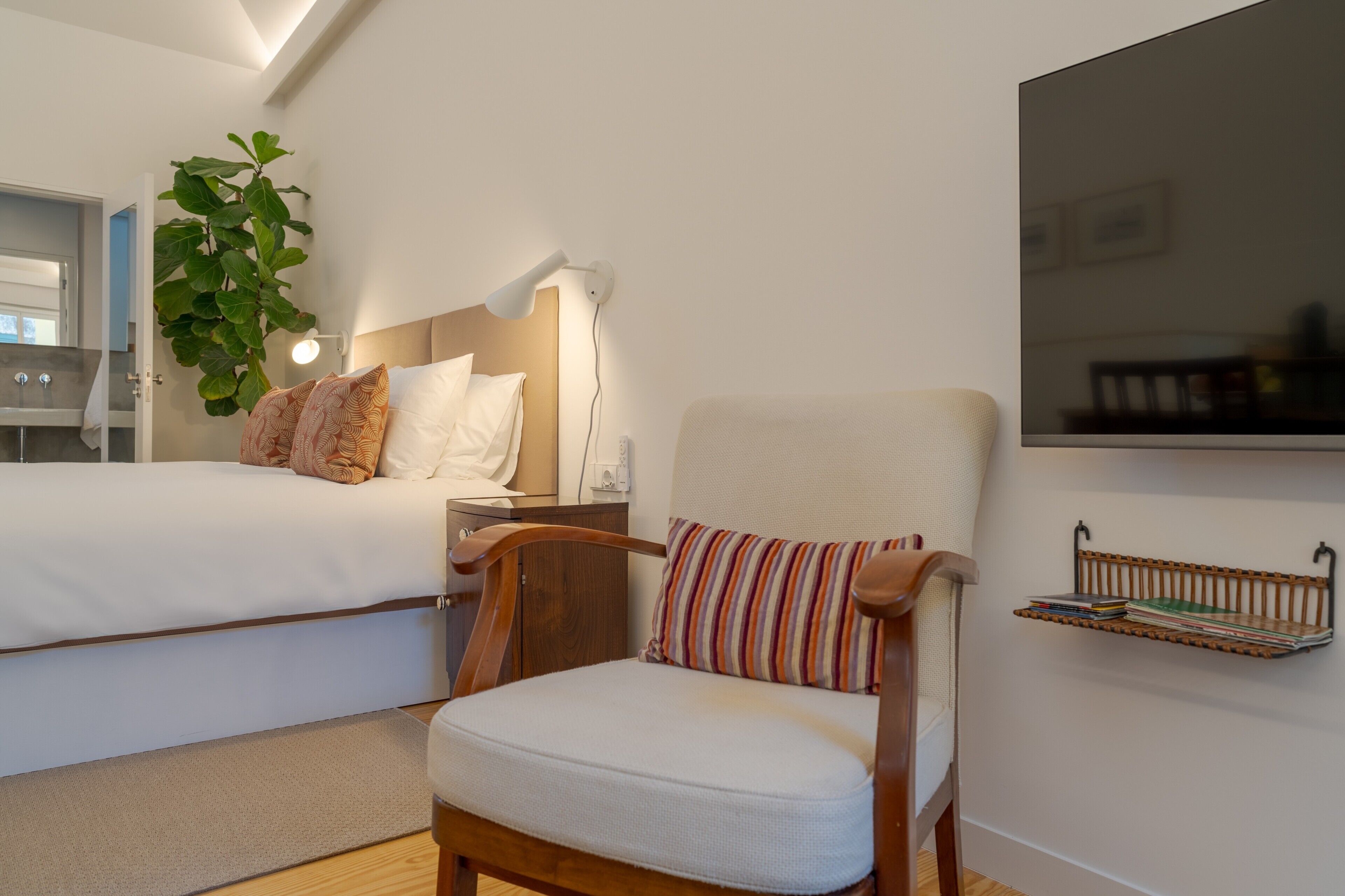 Ein Schlafzimmer mit einem Bett, einem Sessel, einem Nachttisch, einer Lampe, einem an der Wand montierten Fernseher und einer Pflanze.