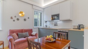 Una cocina compacta con microondas, tostadora y horno, una mesa de comedor con fruta, un sofá rosa y un ventanal.
