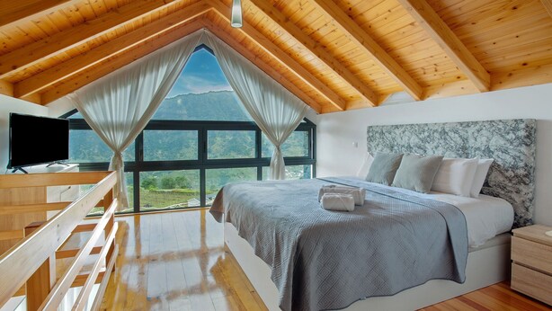 Ein Schlafzimmer mit Holzbalkendecke, einem Bett mit grauer Decke, einem Nachttisch und einem Fernseher auf einem Holzboden.