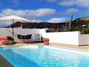 Une piscine avec un mur blanc, une chaise longue rouge, des chaises noires et un parasol blanc.
