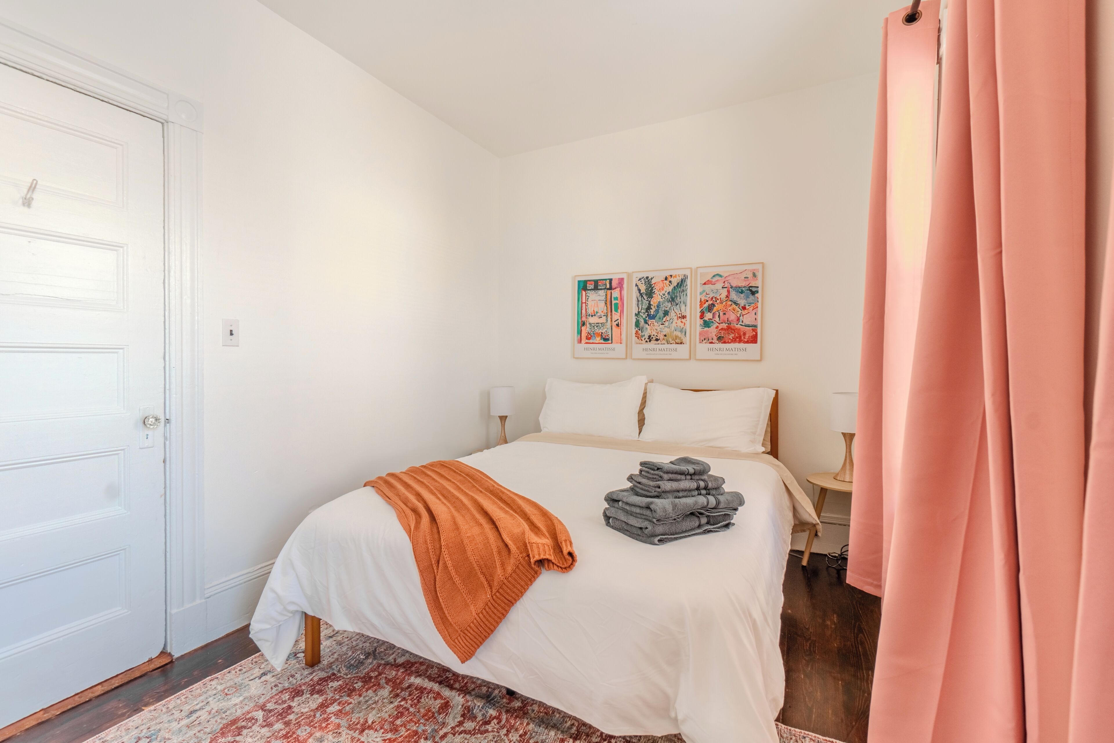 A neatly made bed with a white comforter and an orange throw blanket, a stack of folded towels, and three framed artworks on the wall.