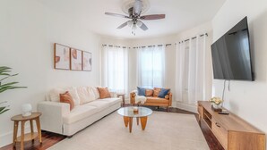 A modern living room with a white sofa, a glass coffee table, a wooden side table, a leather sofa, and a flat-screen TV mounted on the wall. - Spacious 3BDR near Harvard/5-Min Walk to Train - Davis Sq / Free Parking! (Somerville)