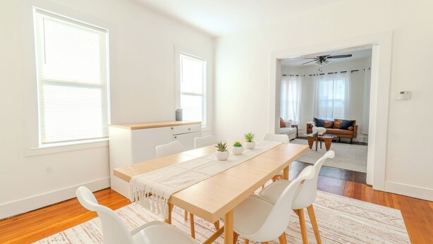 A modern dining area with a wooden table, white chairs, and a kitchen in the background. - Spacious 3BDR near Harvard/5-Min Walk to Train - Davis Sq / Free Parking! (Somerville)