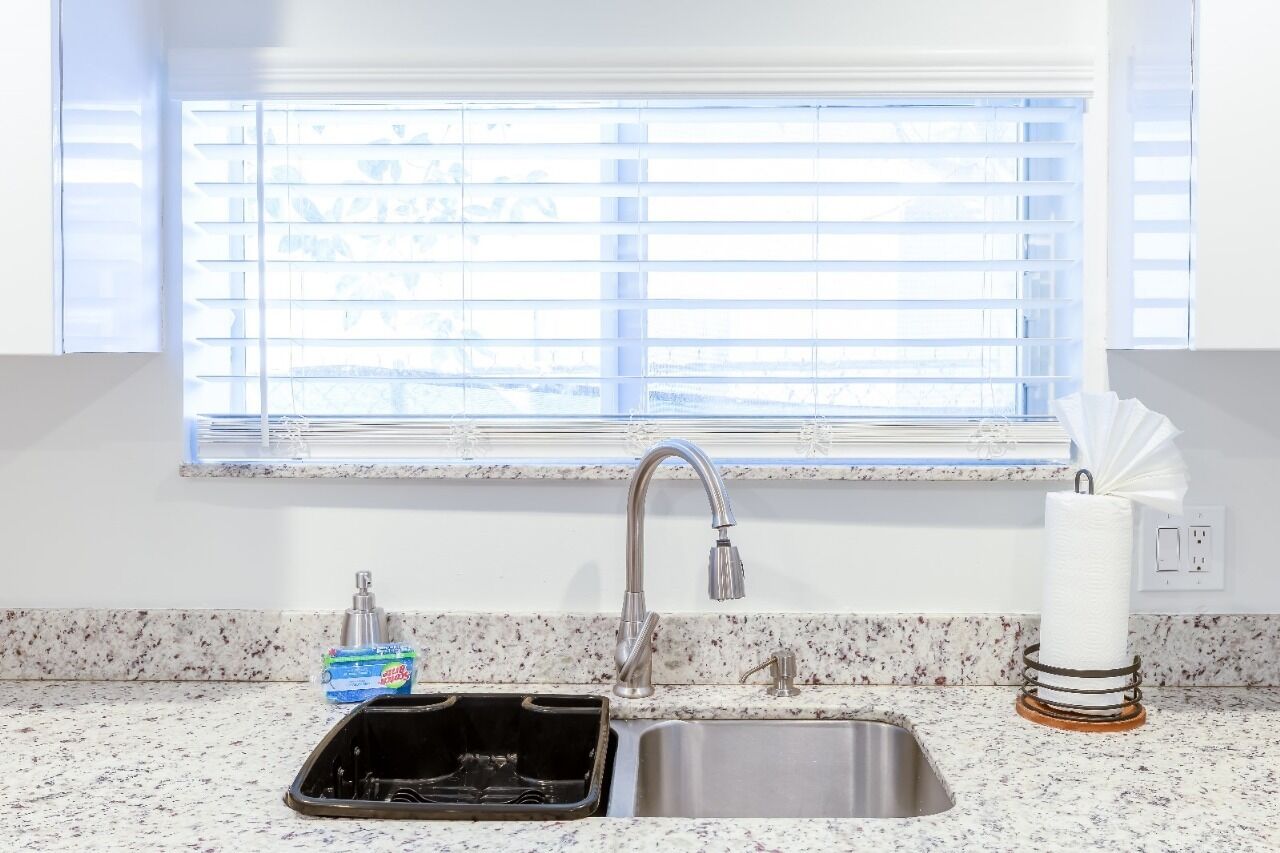 A kitchen with a double sink, a window with blinds, a soap dispenser, and a paper towel holder.