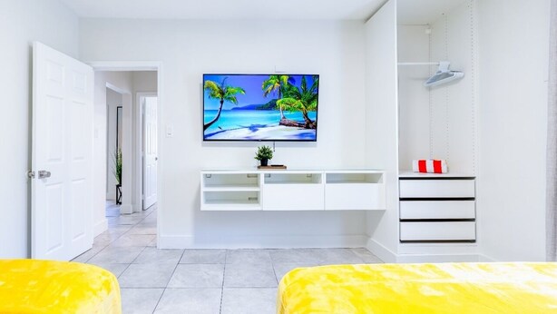 A modern bedroom with a large TV displaying a tropical beach scene, a white wardrobe, and a yellow bed.