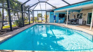 Une piscine entourée de parois en verre, un coin cuisine avec un téléviseur et une terrasse avec des sièges d’extérieur.