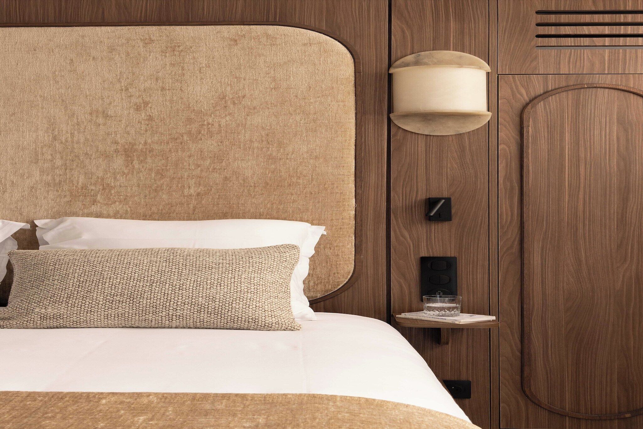 Une chambre d’hôtel moderne avec un grand lit, une tête de lit en bois, une table de chevet avec une lampe et une petite étagère avec un verre.