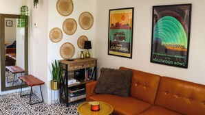 A living room with a brown sofa, a coffee table, a bookshelf, and a wall decorated with woven baskets and a framed poster.
