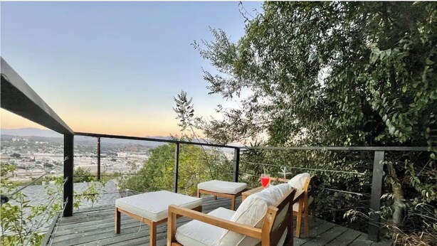 An outdoor deck with wooden furniture, a glass railing, and a view of a cityscape.