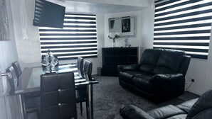 A modern living room with a black leather sofa, a dining table with chairs, and a TV mounted on the wall. - Seaside Stay Sleeps 6 With Pets (Great Yarmouth)