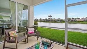 A balcony with wicker chairs, a table with a pitcher and glasses, and a view of a lake with houses in the background. - Sandy Bottoms - Resort Style Community Ammenities, Sandy Bottoms! (Cape Coral)