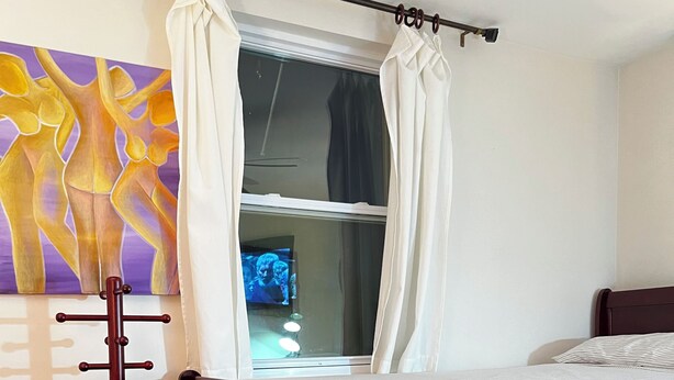 A bedroom with a wooden bed, a window with curtains, a painting on the wall, and a wooden floor.