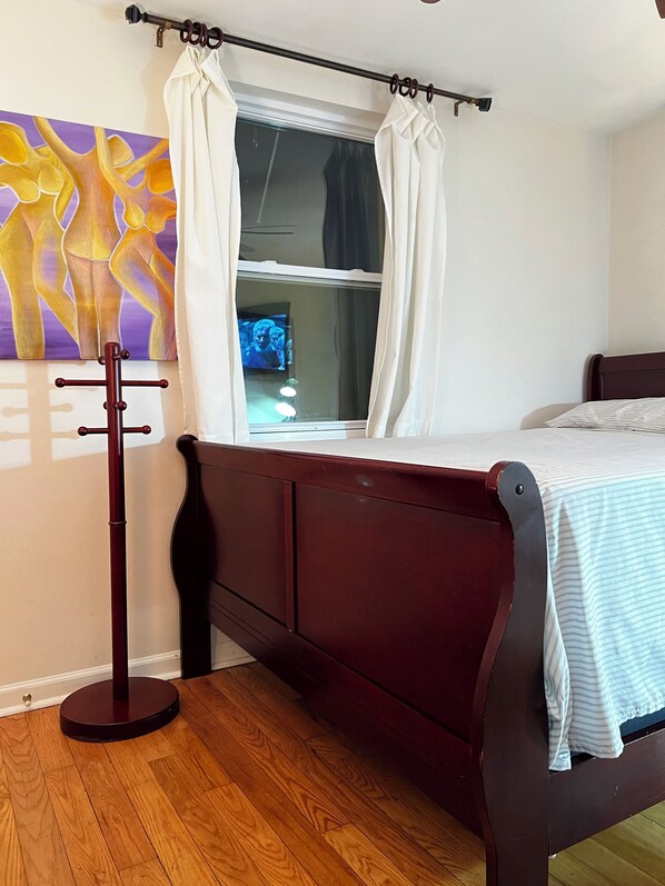 A bedroom with a wooden bed, a window with curtains, a painting on the wall, and a wooden floor.