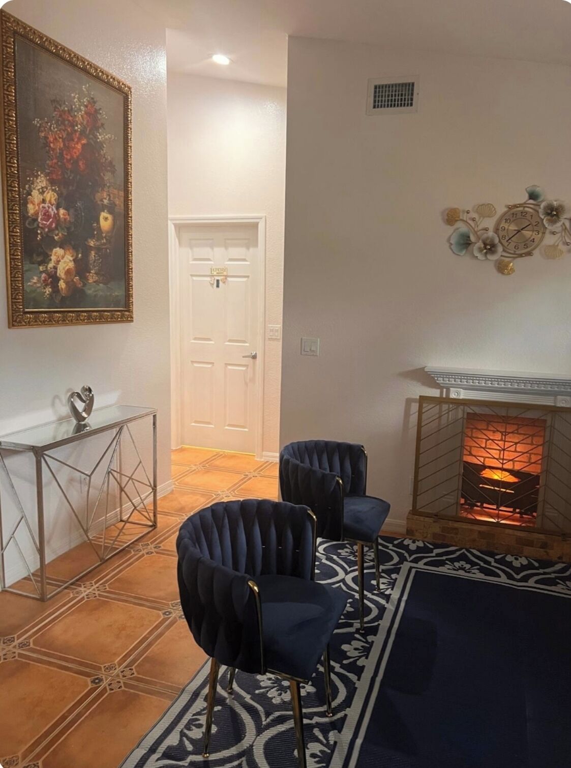Ein Wohnzimmer mit Kamin, zwei dunkelgrauen Sesseln, einem gemusterten Teppich, einem Glastischchen und einem gerahmten Gemälde.