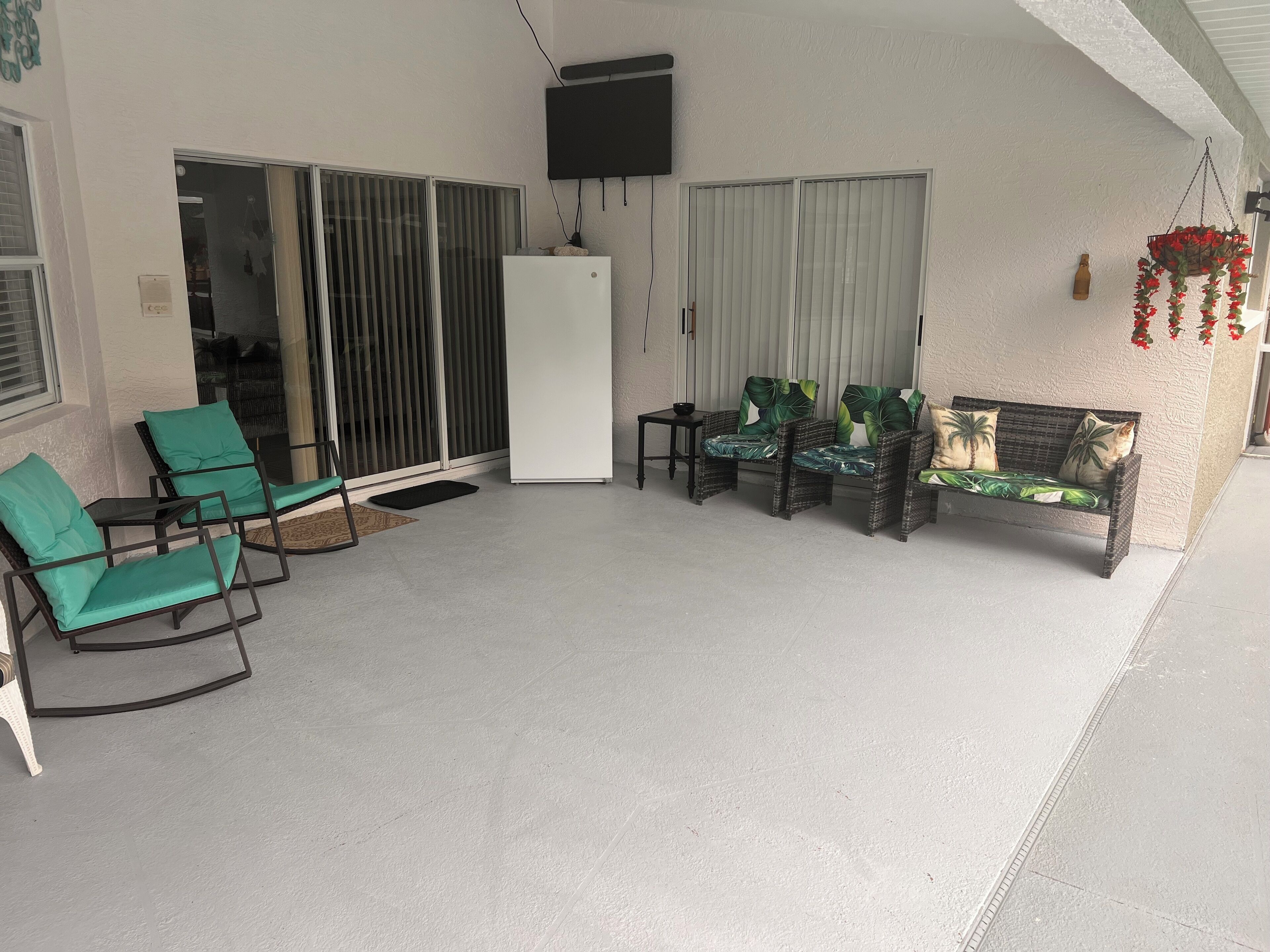 Un espace extérieur abrité, aménagé avec des sièges comprenant des chaises et un canapé, un réfrigérateur et un téléviseur fixé au mur.